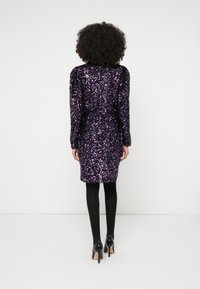 Γυναίκα με σγουρά μαλλιά που φοράει μωβ φόρεμα με παγιέτες, μακρυμάνικο και μήκος μέχρι το γόνατο, μαύρες καλσόν και μαύρες ψηλοτάκουνες γόβες, στέκεται μπροστά σε λευκό τοίχο.