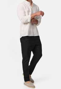 Man in een opgerolde lange mouw witte shirt, zwarte tapered broek en beige instap espadrilles staat tegen een witte achtergrond.