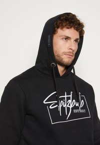 Schwarzer Kapuzenpullover mit Kordelzug, der ein auffälliges weißes Logo-Design zeigt, das in einem rechteckigen Rahmen "Spitzbulb EST. 2017" liest. Weiche Stoffoberfläche.