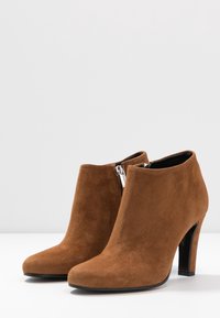 Brune ruskindsankelstøvler med spids tå, høj hæl og lynlås i siden. Glat tekstur med et rent design og få accenter.
