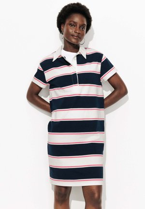 Femme avec des cheveux bouclés courts portant une robe polo à manches courtes rayée navy, blanche et rose, se tenant debout avec les mains derrière le dos.