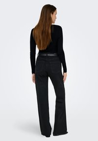Femme aux longs cheveux bruns portant un haut à manches longues noir, un pantalon large noir taille haute et des chaussures noires à clous, se tenant de dos.