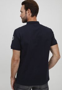 Mørkeblå poloskjorte laget av bomull. Har krage, korte ermer og et logo-patch på venstre arm. Designet for en avslappet passform.