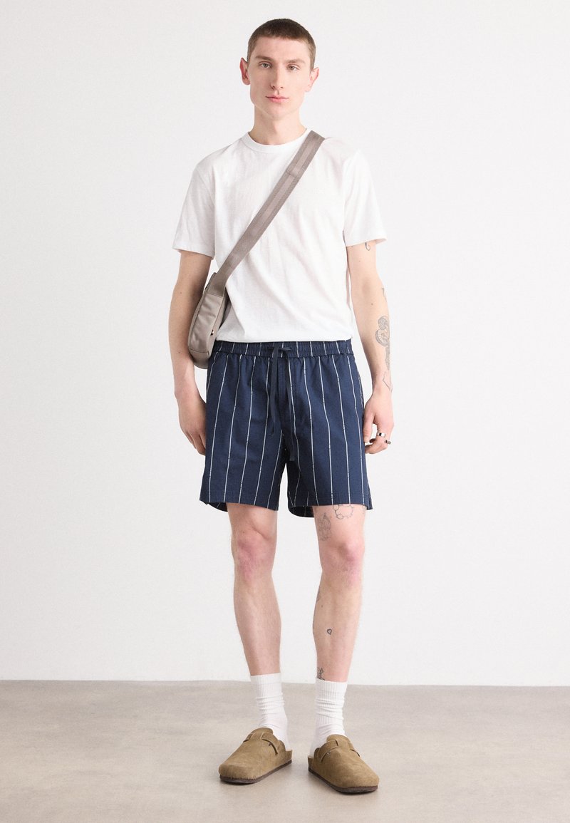 Marineblau- und weiß gestreifte Shorts mit elastischem Bund, kombiniert mit einem schlichten weißen T-Shirt und beigen Slippern. Kleine Umhängetasche.