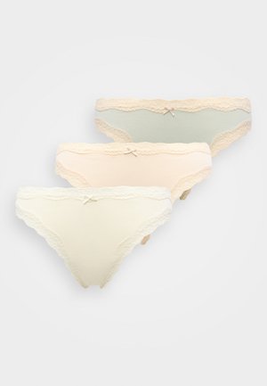 Femme aux cheveux bruns courts portant un soutien-gorge couleur chair et une culotte beige bordée de dentelle, debout devant un fond clair uni.