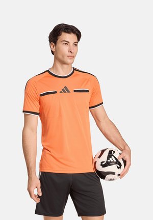 Junger Mann in orange-schwarzem Fußballtrikot, der auf weißem Hintergrund einen schwarz-weißen Adidas-Fußball hält.