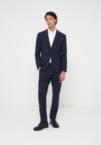 Μαύρο κοστούμι που περιλαμβάνει ένα εφαρμοστό σακάκι με ένα μόνο κουμπί, παντελόνι σε ίδια απόχρωση και λευκό πουκάμισο, συμπληρωμένο με μαύρα κοσμικά παπούτσια.