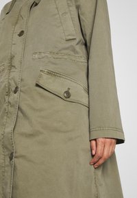 Oliwkowa kurtka bawełniana o luźnym kroju, z boczną kieszenią naszytą i dużymi guzikami z przodu. Obecne detale szwów.