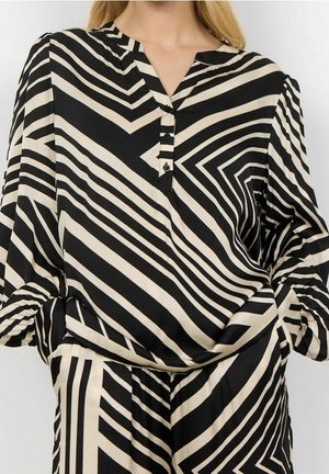 Femme portant un chemisier à manches longues rayé géométrique noir et beige avec trois boutons et un pantalon assorti, les mains dans les poches.