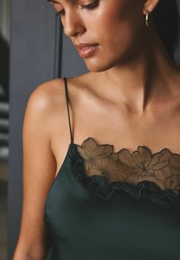 Dunkelgrünes Satin-Camisole mit floralen Spitzenbesätzen am Ausschnitt, dünnen Trägern und glatter Textur.
