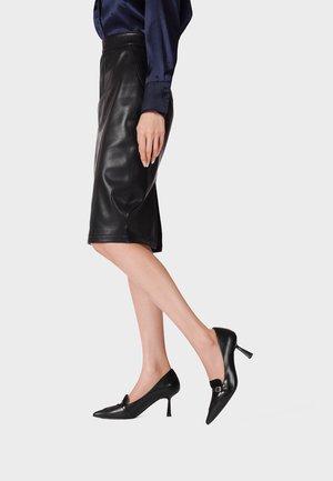 Donna che indossa una gonna in pelle nera al ginocchio e scarpe nere con tacco alto, con un piede leggermente sollevato, in piedi davanti a uno sfondo chiaro.