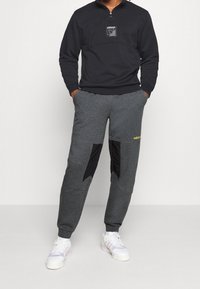 Svart zippartröja med logotyp på bröstet, tillsammans med mörkgrå sweatpants med svarta och gula detaljer samt sidfickor.