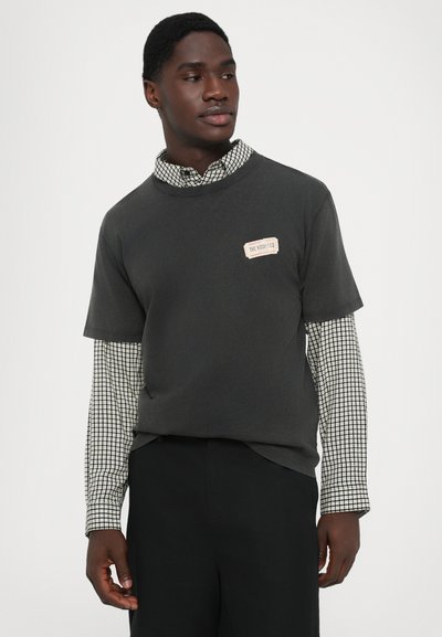 Homem vestindo uma t-shirt preta de manga curta por cima de uma camisa de manga longa com padrão, combinado com calças pretas, olhando para o lado.