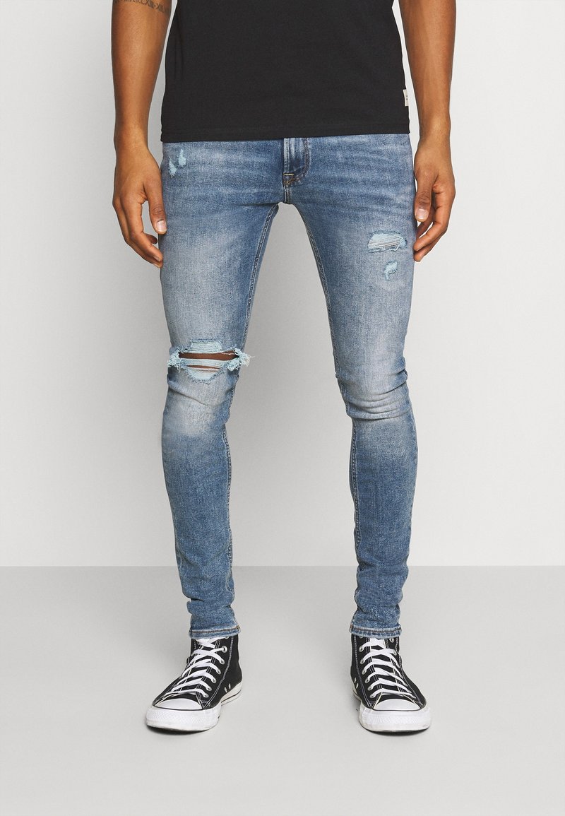Tajta blå denimjeans med reva detaljer vid knäna, ljus blekning och en lätt textur, kombinerat med svarta högsnörade sneakers.