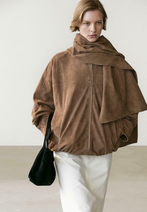 Donna che indossa una giacca oversize in pelle scamosciata marrone con dettaglio sciarpa, pantaloni bianchi e tiene una borsa nera su sfondo semplice.