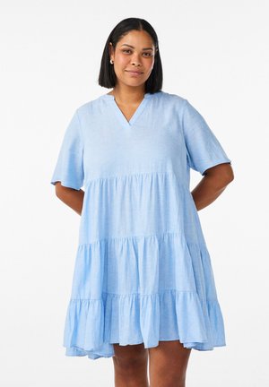 Žena s krátkymi tmavými vlasmi v širokých svetlomodrých vrstvených šatách s krátkymi rukávmi, stojaca s rukami za chrbtom.