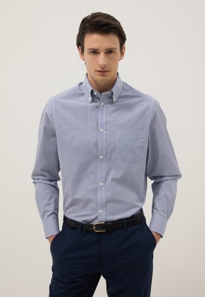 Giovane uomo in piedi con le mani nelle tasche, indossa una camicia a quadri azzurro chiaro con bottoni e pantaloni blu scuro con cintura nera.