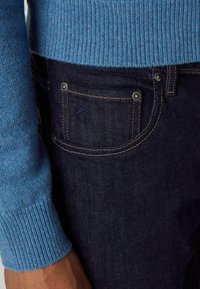 Detailný záber osoby nosiacej modrý pletený sveter a tmavomodré rifľové nohavice s viditeľnými švami a kovovými nitmi na vrecku.