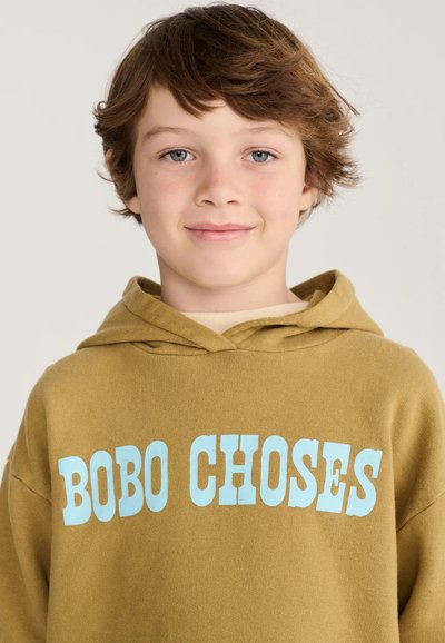 Jeune garçon aux cheveux châtain clair portant un sweat à capuche moutarde avec un texte bleu "BOBO CHOSES" sur un fond clair.