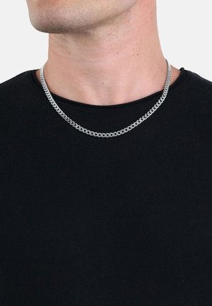 Collana in argento con maglie intrecciate, indossata su una maglietta nera a girocollo. Presenta una superficie liscia e lucida.