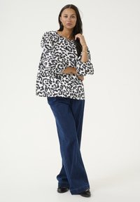 Haut à motifs léopard en blanc et noir, avec une coupe décontractée et des manches longues, associé à un jean large bleu foncé et des chaussures noires.