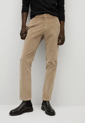 Mann trägt beige Cordhosen, schwarzes Langarmshirt und schwarze Lederstiefeletten, steht vor einem einfarbigen grauen Hintergrund.
