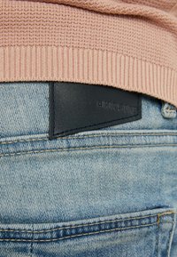 Ljusblå denimjeans med en texturerad yta och en mörkblå läderlapp vid midjan, med det präglade varumärket "CHASING."