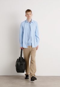 Lys blå knapp-up skjorte over en lys blå topp, kombinert med beige bukser og svarte sko, bærende en svart myk tote bag.