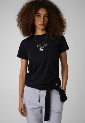 Donna con capelli ricci che indossa una t-shirt nera Guess, pantaloni della tuta grigi e collane a strati, in piedi su uno sfondo grigio semplice.