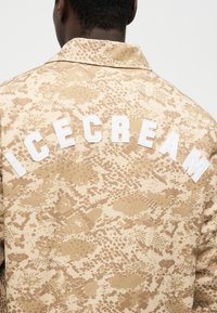 Asmenų atgalinis vaizdas, dėvintis smėlio spalvos raštuotą švarką su dideliais baltais užrašais „ICECREAM“ viršutinėje nugaros dalyje.