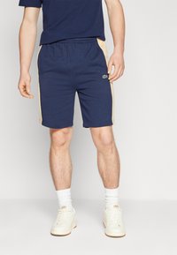 Shorts bleu marine avec des bandes latérales beiges, taille élastique et un logo vert. Portés avec des chaussettes blanches et des baskets claires.