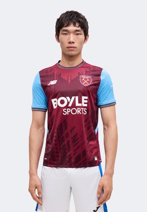 Jeune homme portant le maillot de football West Ham United bordeaux et bleu avec le logo Boyle Sports et un short de sport blanc, debout devant un fond uni.
