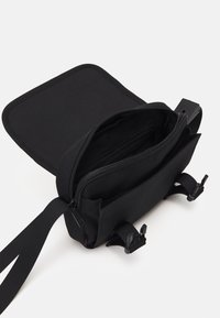 Borsa a tracolla in tessuto nero con pattina aperta che rivela un compartimento principale e una tasca con zip. Cinghie regolabili e accessori in plastica.