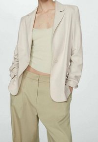Světle béžové sako na míru s jedním knoflíkem, doplněné o žebrovaný světle zelený top na ramínka a kalhoty s vysokým pasem v khaki barvě. Hladká textura.