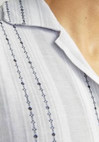 Chemise blanche avec col, ornée de motifs brodés verticaux bleus et d'un tissu texturé, mettant en valeur des accents bleus le long des lignes.