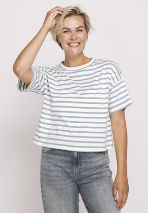 Lächelnde Frau mit kurzen blonden Haaren, die ein weißes und hellblau gestreiftes Hemd und hellgraue Jeans trägt und sich an den Haaren berührt.