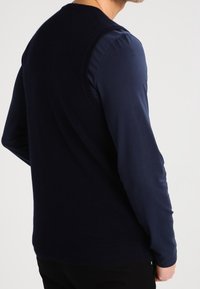 Marinblå ärmlös tröja över en långärmad skjorta; mjuk textur; ribbad stickning vid hals och ärmhål; lagerdesign.