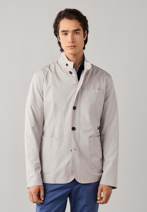Jeune homme portant une veste boutonnée gris clair avec un col montant et un pantalon bleu, debout devant un fond uni.