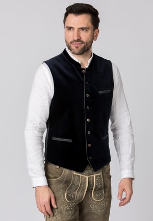 Dunkelblaues Samtwesten mit hohem Kragen, vorderer Knopfverschluss, kontrastierenden grauen Nähten und zwei kleinen Taschen. Kombiniert mit einem weißen Hemd.