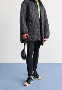 Chaqueta acolchada negra con acolchado en rombos y cuello alto, top verde corto, leggins negros y zapatillas negras con rayas en teal y acentos blancos.