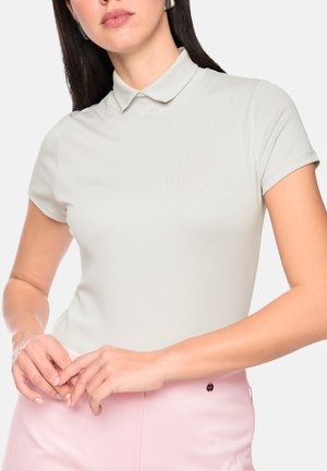 Woman with long dark hair wears light gray short-sleeve polo shirt and light pink pants, hands loosely clasped at waist.