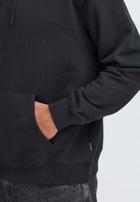 Svart hettegenser med ribbestrikkede ermer og nederkant, med en frontkengurulomme og subtil merking på siden. Materialet er en bomullsblanding.