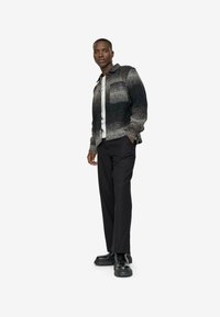 Camisa de lana a cuadros gris y negro con un bolsillo frontal, combinada con pantalones negros y zapatos negros gruesos, de pie frente a un fondo blanco.