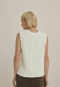 Pull sans manches blanc avec un col et des emmanchures côtelés, texture douce et ourlet arrière légèrement plus long. Aucun motif ou élément en métal visible.