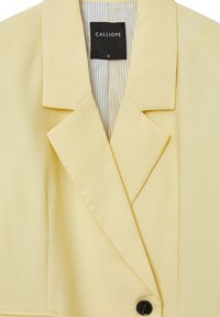 Blazer giallo con colletto strutturato, chiusura a singolo bottone nero e fodera plissettata. Realizzato in un tessuto liscio con una leggera lucentezza.