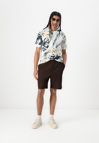 Camisa tropical de manga corta con base blanca y patrones de hojas verdes y amarillas, combinada con pantalones cortos marrones con pliegues y zapatillas crema.