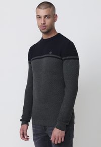 Jersey de punto con un cuerpo texturizado de color gris oscuro, parte superior de color azul marino sólido y un pequeño logo en el pecho. Cuello redondo y puños de canalé.