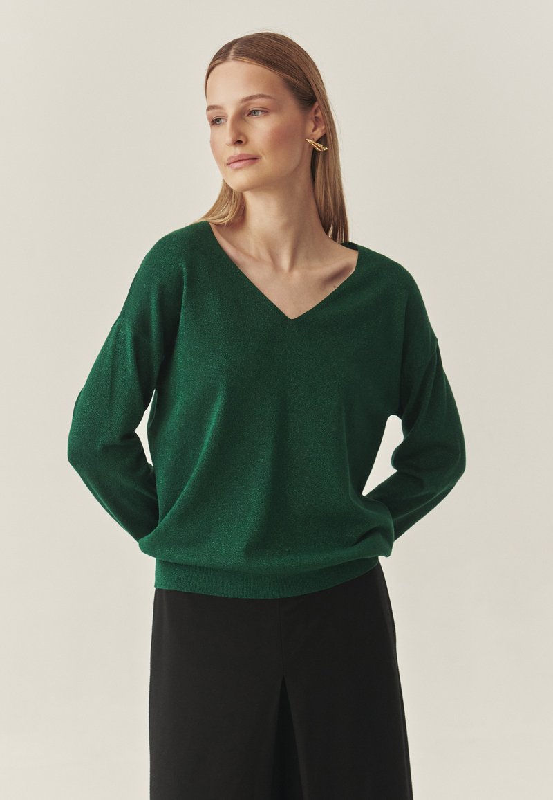 Zaļš, mirdzošs adīts džemperis ar V veida izgriezumu, vaļējām garām piedurknēm un ribota auduma apakšu, kombinēts ar melnu svārku.