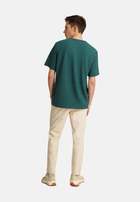 Tyrkysové tričko s krátkým rukávem s texturovaným vzorem, kombinované s krémovými kalhotami a světlými teniskami. Jednoduchý, ležérní design z pohledu zezadu.