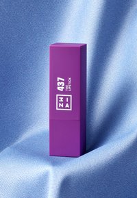Étui à rouge à lèvres rectangulaire violet avec du texte blanc indiquant "ZINA 437 LE ROUGE À LÈVRES." Mis en valeur sur un fond bleu texturé.